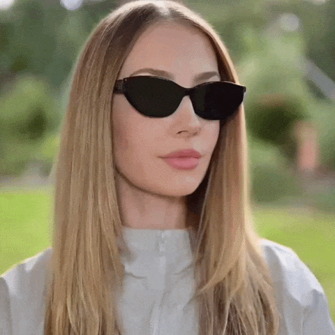 A woman with long blonde hair wearing black cat-eye sunglasses and a light gray zip-up jacket, photographed outdoors with a blurred green garden background. She is turning her head left and right to showcase the sunglasses.