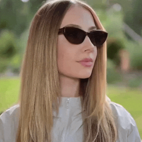A woman with long blonde hair wearing brown cat-eye sunglasses and a light gray zip-up jacket, photographed outdoors with a blurred green garden background. She is turning her head left and right to showcase the sunglasses.