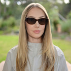 A woman with long blonde hair wearing dark brown cat-eye sunglasses and a light gray zip-up jacket, photographed outdoors with a blurred green garden background.