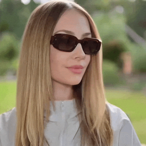 A GIF of a woman with long blonde hair wearing dark brown cat-eye sunglasses and a light gray zip-up jacket, photographed outdoors with a blurred green garden background. She is turning her head left and right to showcase the sunglasses.