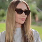 A woman with long blonde hair wearing cat-eye sunglasses with dark brown frames and dark lenses, set against a blurred green outdoor background. She is turning her head left and right to showcase the sunglasses.