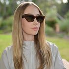 A woman with long blonde hair wearing cat-eye sunglasses with dark brown frames and dark lenses, set against a blurred green outdoor background.
