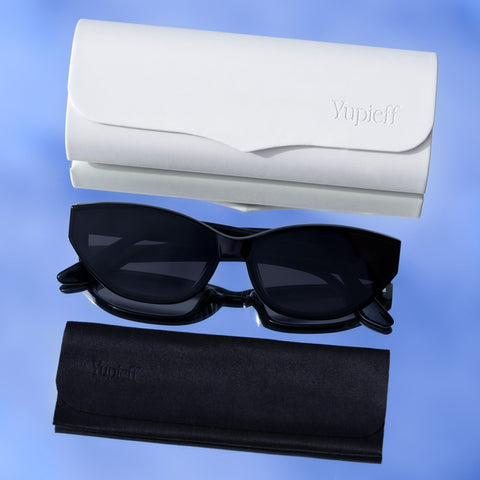 Black cat-eye sunglasses with dark lenses, shown with a white hard case and a black soft case on a blue gradient background. The embossed brand name 'Yupieff' is visible on both cases.